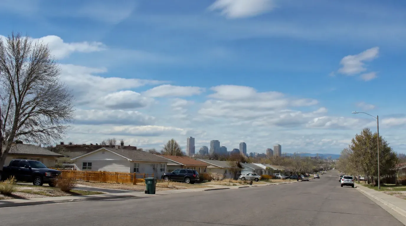 A Commerce City street of modest ranch homes under a wide sky with the Denver skyline far off